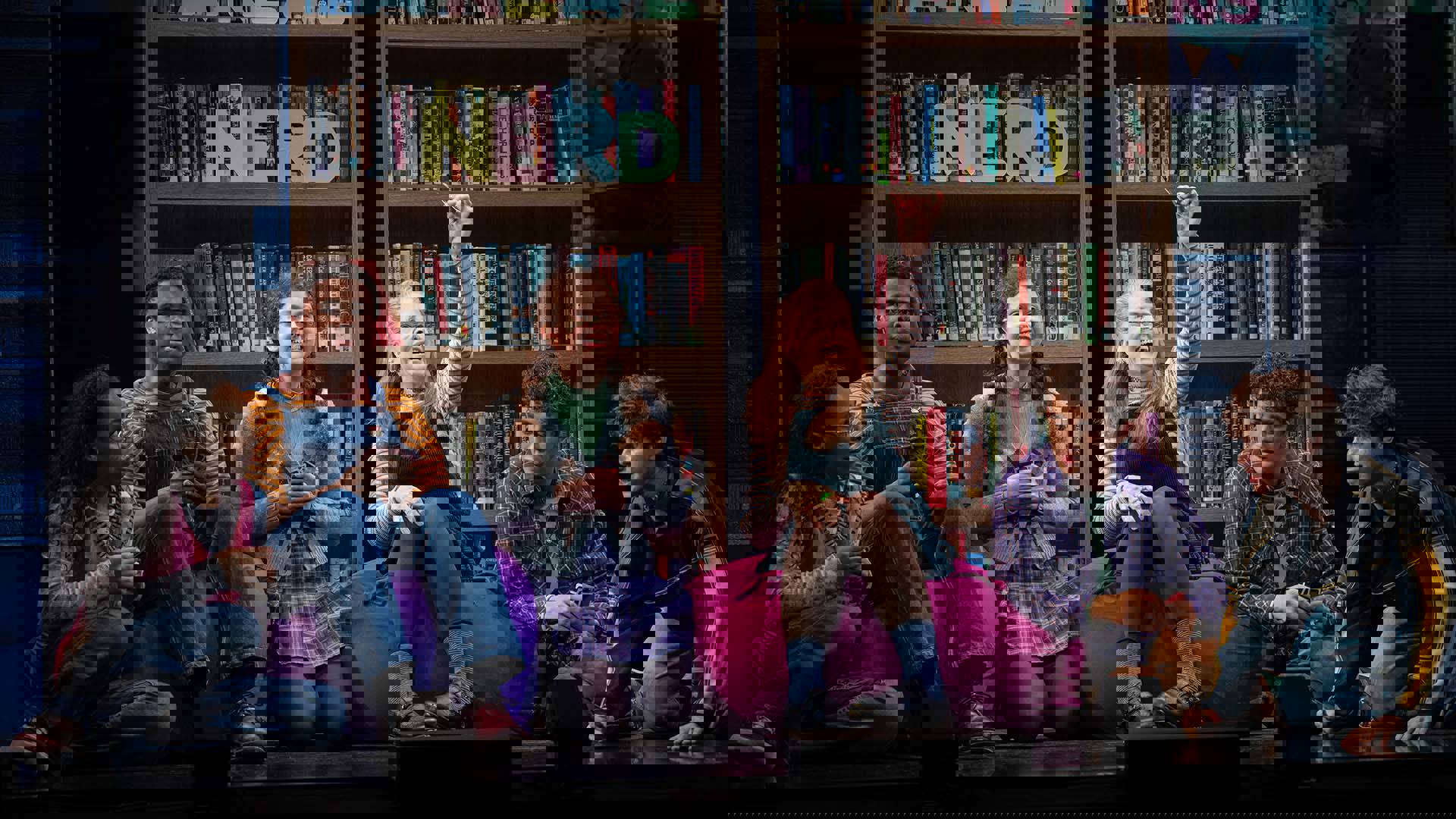 Group of six cast members with diverse skin tones sit on bean bags in front of two large bookshelves.