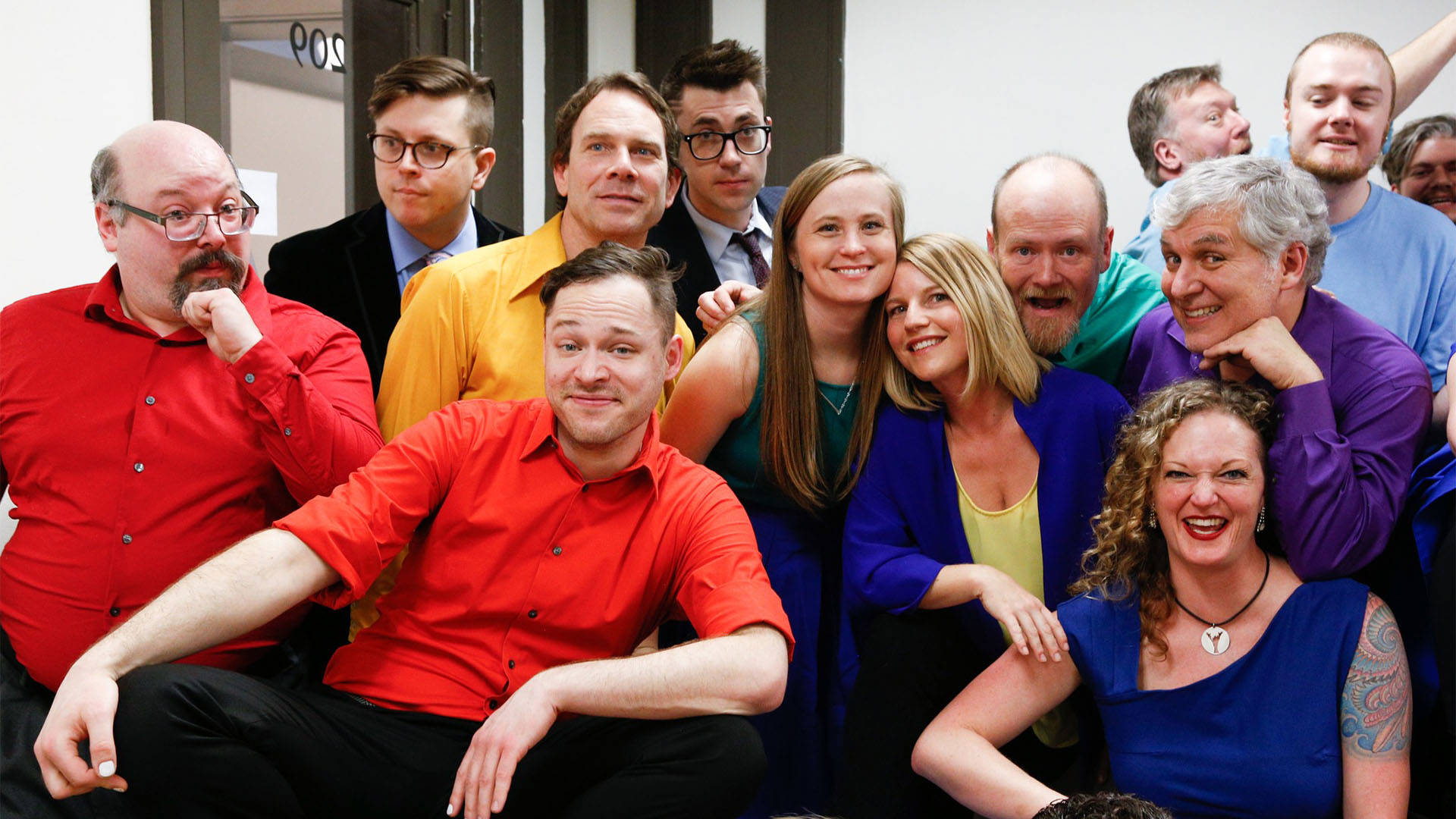 A group of people wearing vibrant colored dress clothes in an office space, some are sitting down with others crowding behind them. Facing the camera, many are making a variety of silly and smiling faces.