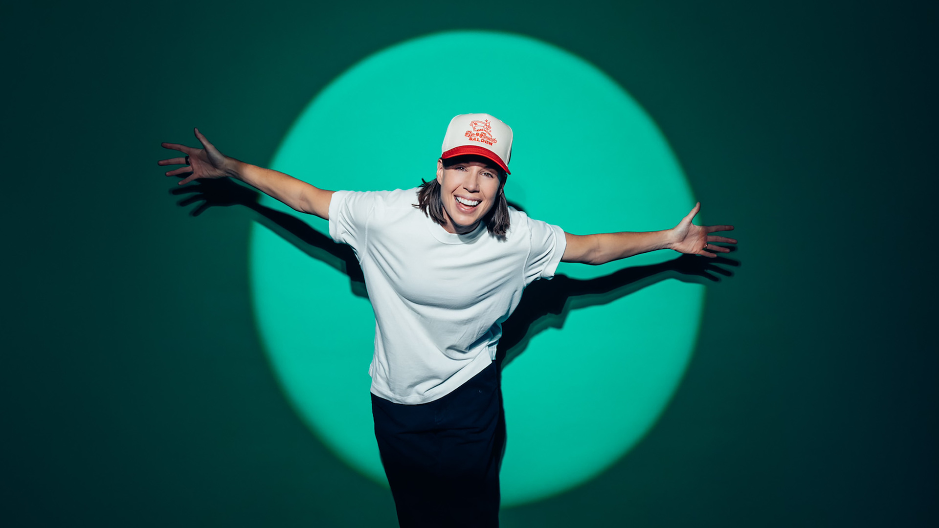 Photo of a smiling Danae Hays with arms outstretched against a green wall with a spotlight. She has light complexion, medium length brown hair, a white shirt, dark pants and a trucker hat.