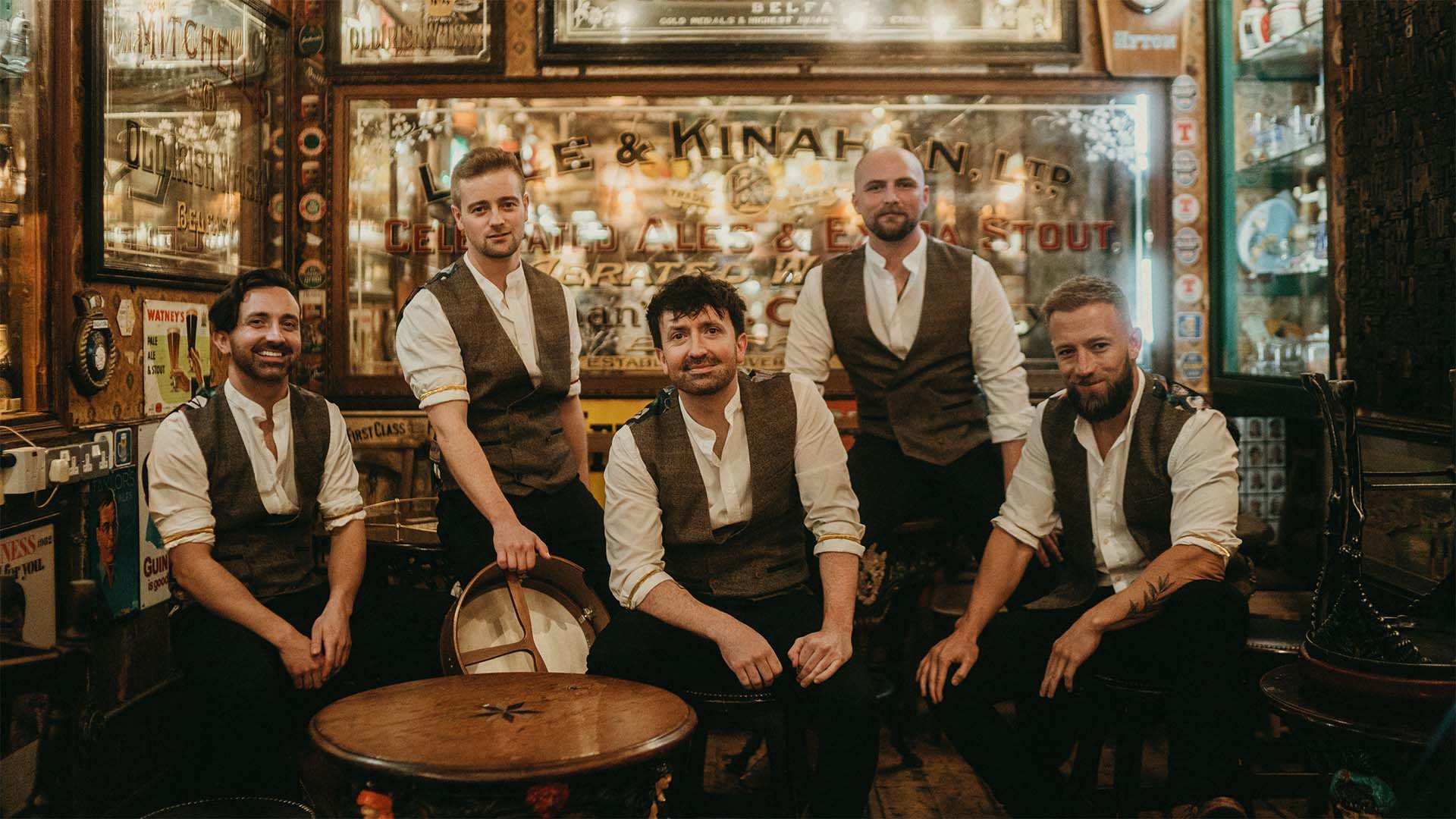 Five people with various forms of facial hair wearing dark pants, white shirts and brown vests sit in an Irish themed bar. 