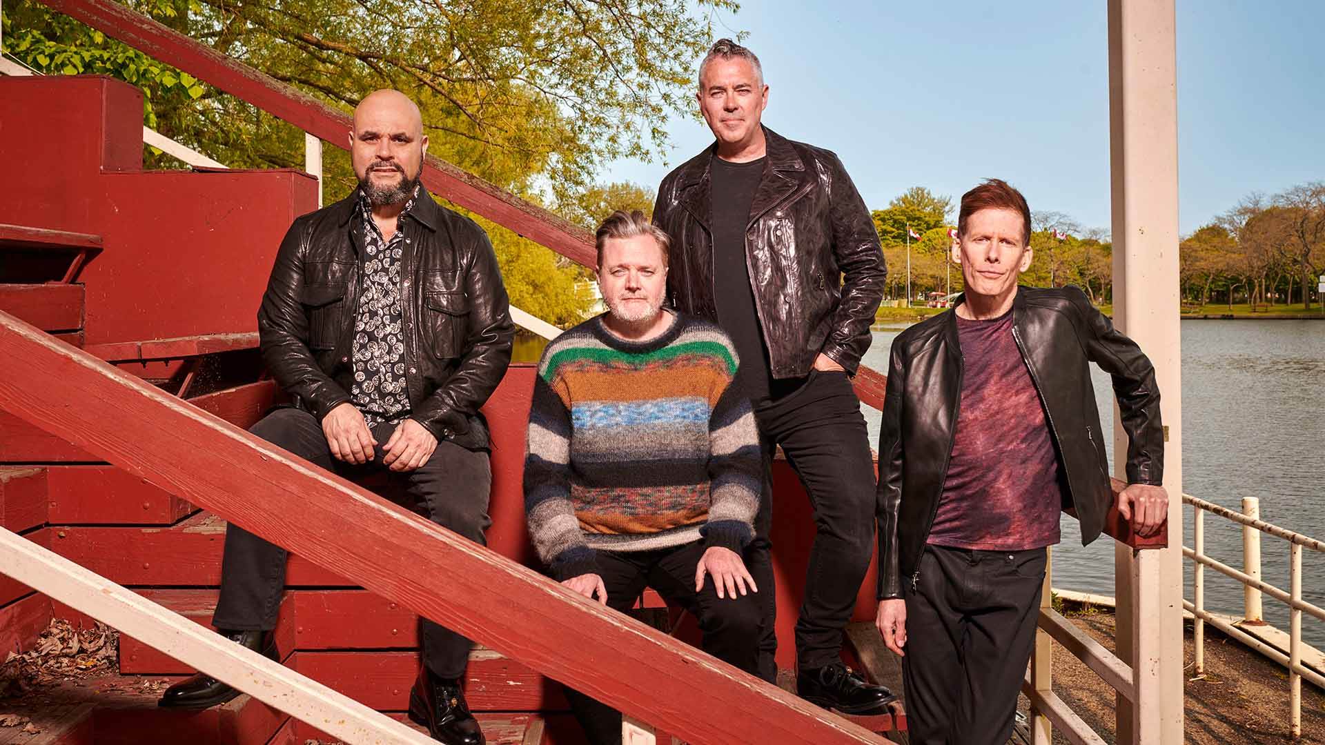Barenaked Ladies band - four middle-aged men sitting and leaning against red wooden bleachers that overlook a lake with trees. Three men are wearing black leather jackets and black jeans with dark shirts. In the center, a man with graying short hair wearing a black, brown and green striped sweater.