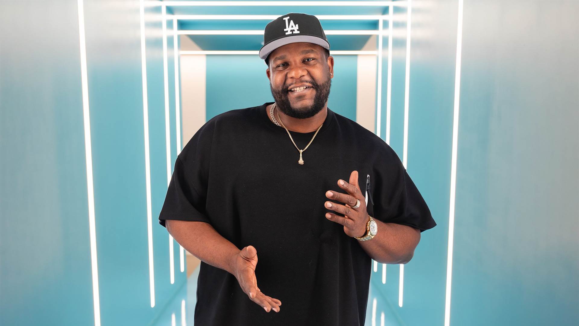 Nate Jackson, a black man with a light beard wearing a black t-shirt, gold chain and black billed cap with the LA Dodgers "LA" on it. He is smiling and standing in a light blue hallway with white lights.